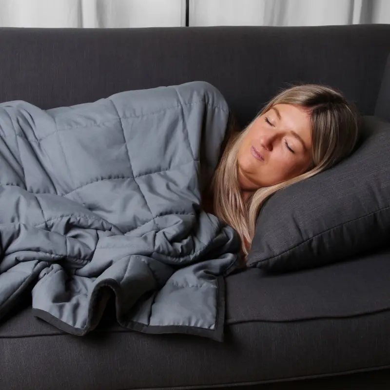Donna riposa su un divano coperta dalla yourGEAR Como coperta ponderata 6 kg in cotone grigio, trapuntata, dimensioni 135x200 cm, ideale per terapia con pressione profonda e rilassamento.
