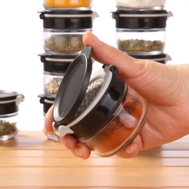 Hand holding a your GEAR Double-Flip spice jar with black lid and built-in shaker, filled with spices; several similar glass spice jars are stacked in the background, highlighting kitchen organization.