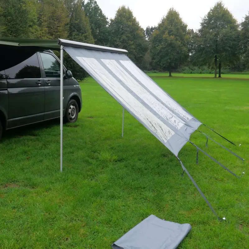 Paroi avant your GEAR Panorama Wall 2,4 x 2 m fixée à une marquise de van, avec fenêtres transparentes pour protection contre soleil, vent et pluie, installée sur pelouse verte en extérieur.