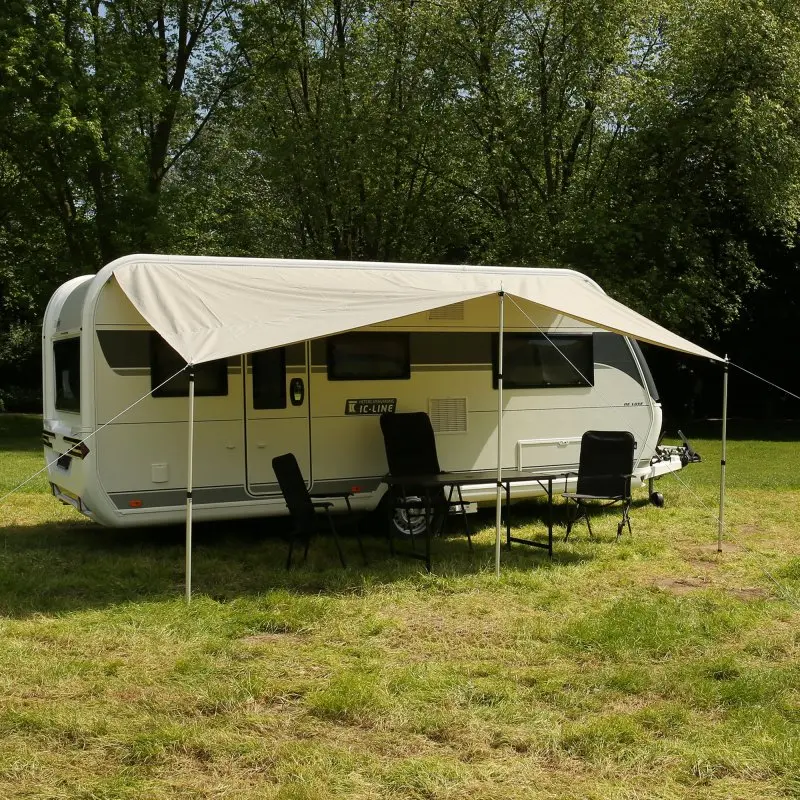 Wohnwagen mit your GEAR Caravan Tarp TC 450 x 240 Baumwoll-Sonnendach, befestigt mit drei Alu-Teleskopstangen, spendet großflächigen Schatten. Outdoor-Sitzgruppe unter dem Sonnensegel auf Wiese.