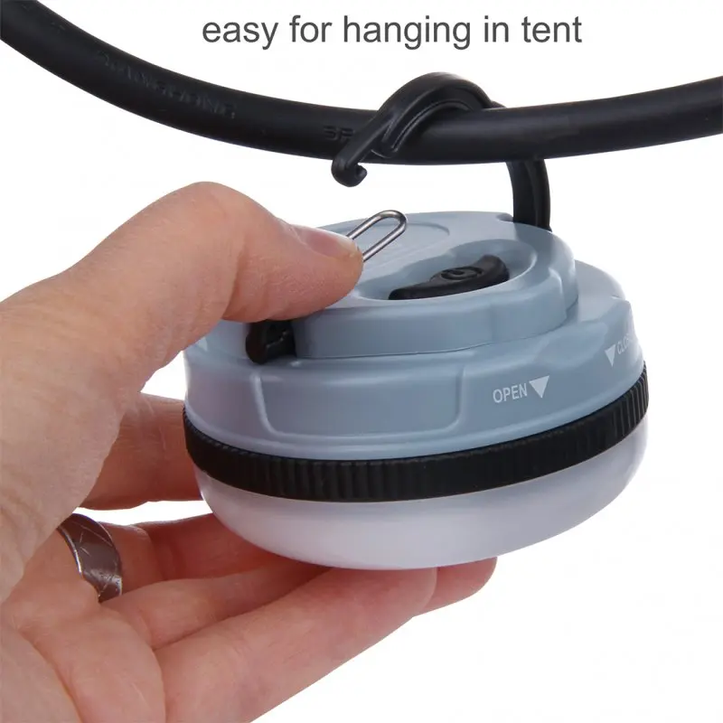 Compact blue 10T MCL 150 camping lamp with 150 lumens, round ABS housing, visible open/close mark, metal hook for hanging, shown attached to a tent pole. Ideal for outdoor lighting.