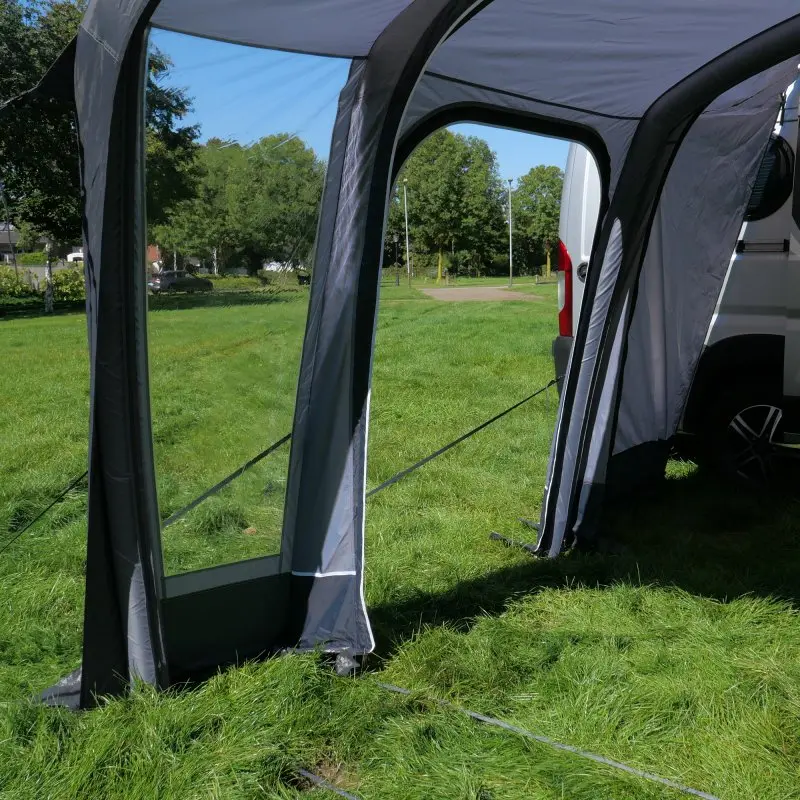 Avance de un toldo inflable your GEAR Manarola Air para furgoneta, con ventanas grandes y puerta lateral, anclado a un vehículo en un área verde, ideal para camping y autocaravanas.