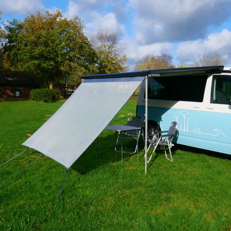Vordere Sonnenschutzwand your GEAR Capri Sun Blocker 2,4 x 2 m an Wohnmobil-Markise montiert, bietet Schatten und Sichtschutz, hellgraues Spezialgewebe, Campingmöbel darunter auf grünem Rasen.