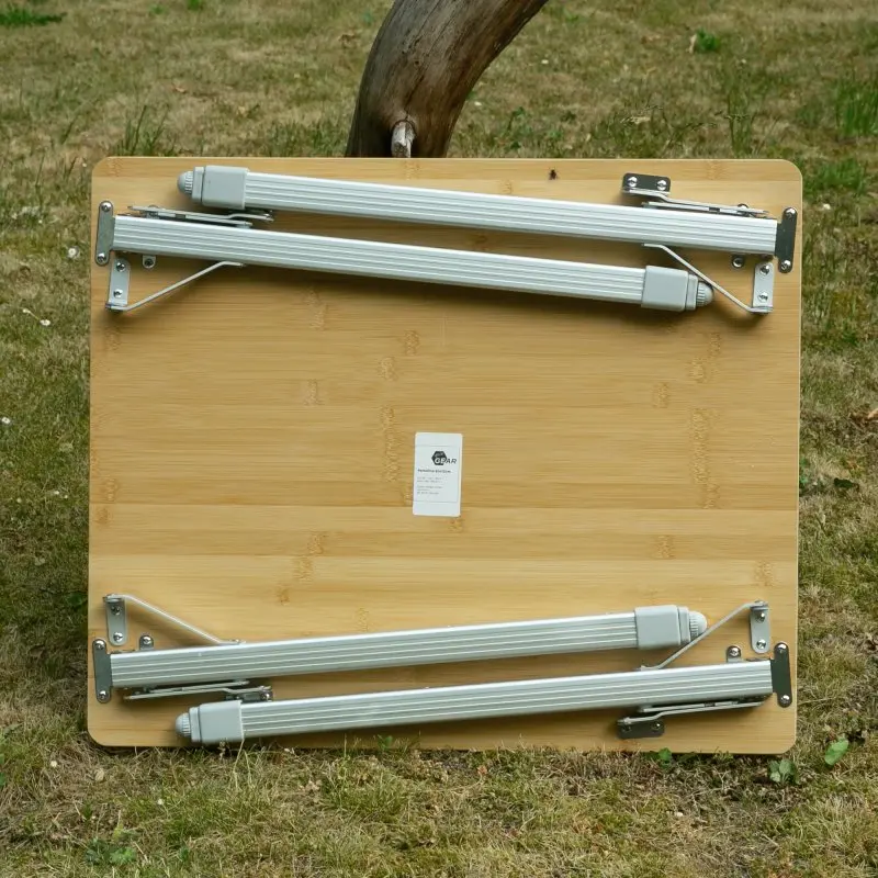 Bambus Campingtisch your GEAR Portofino M mit klappbaren Aluminium-Teleskopbeinen, zusammengeklappt auf Rasen stehend; robuste, glatte Bambusplatte, sichtbare Scharniere und Sicherungen, rechteckige Form.