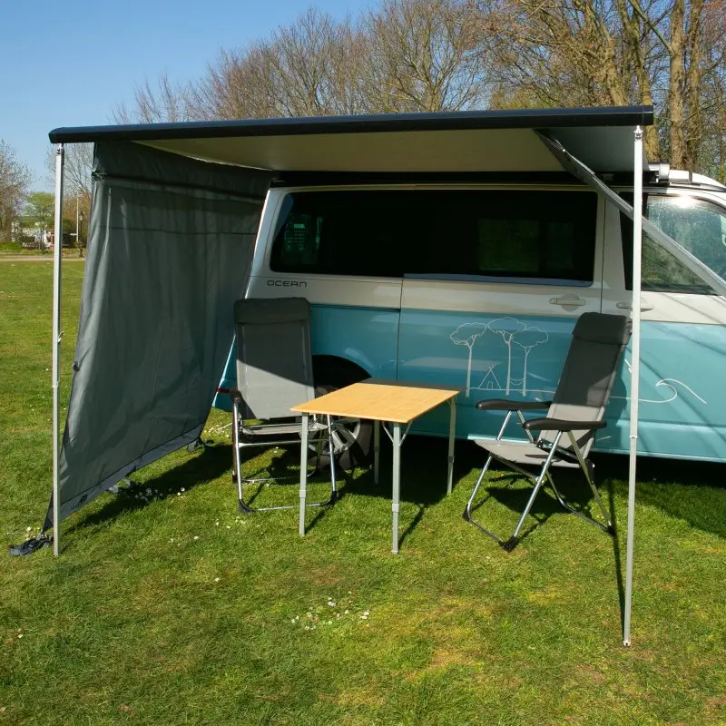 Camping van with your GEAR Sun + Rain Blocker side wall attached to the awning, providing sun and rain protection; includes two chairs and a table under the shelter, ideal for outdoor camping.