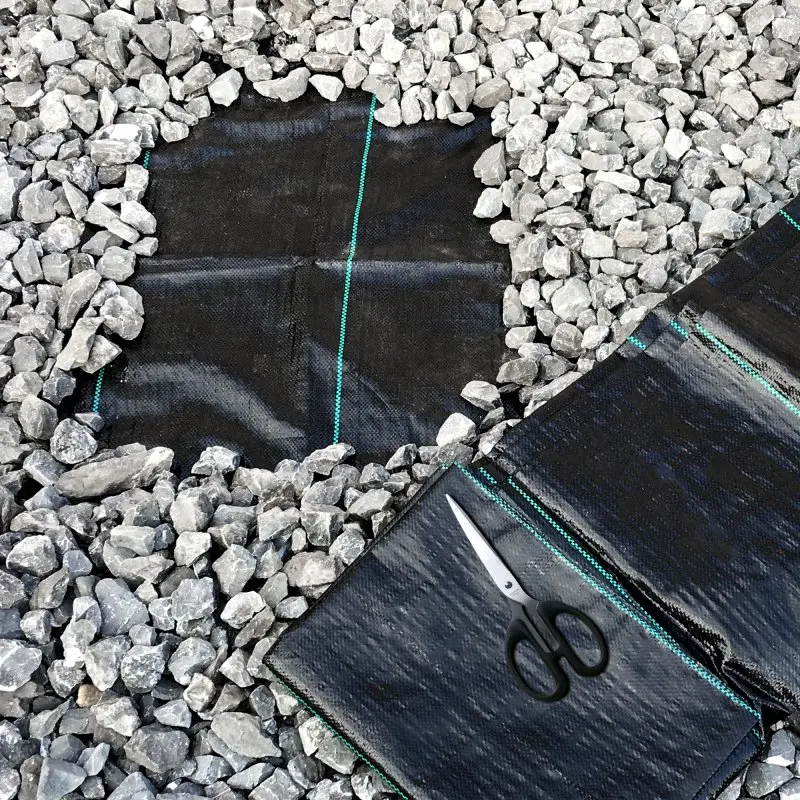 Unkrautfolie yourGEAR 10 m², schwarz mit grünen Linien, zugeschnitten auf Kiesboden, daneben Schere. Reißfestes, wasserdurchlässiges und UV-stabiles Gartenvlies zum Unkrautschutz.