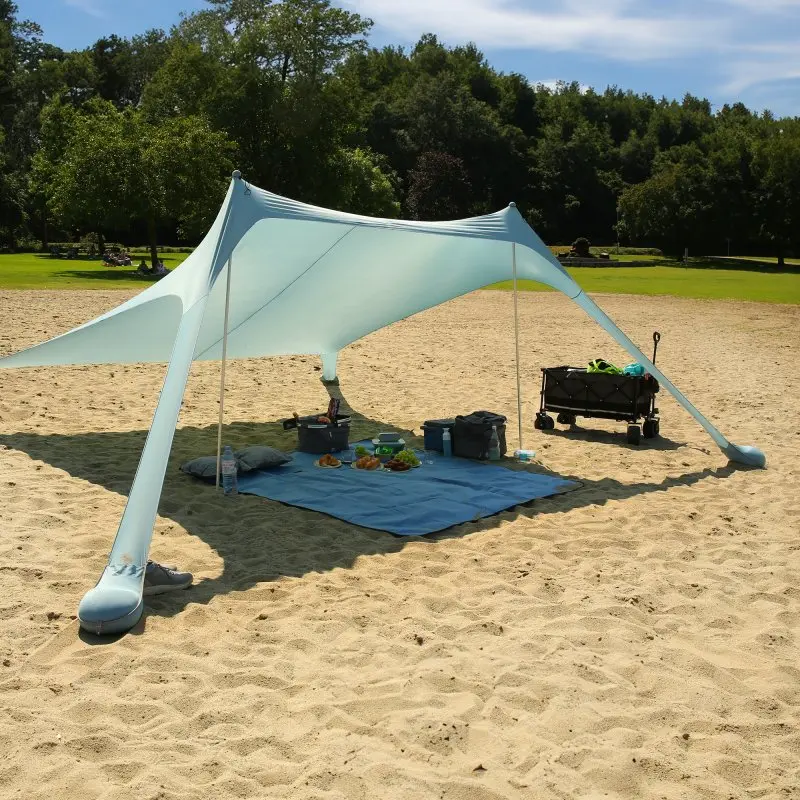 Abri de plage XXL your GEAR Cameo 3x3m en lycra bleu clair, monté sur le sable avec tapis, pique-nique et accessoires dessous, offrant une protection solaire UV 50+ dans un environnement de plein air verdoyant.
