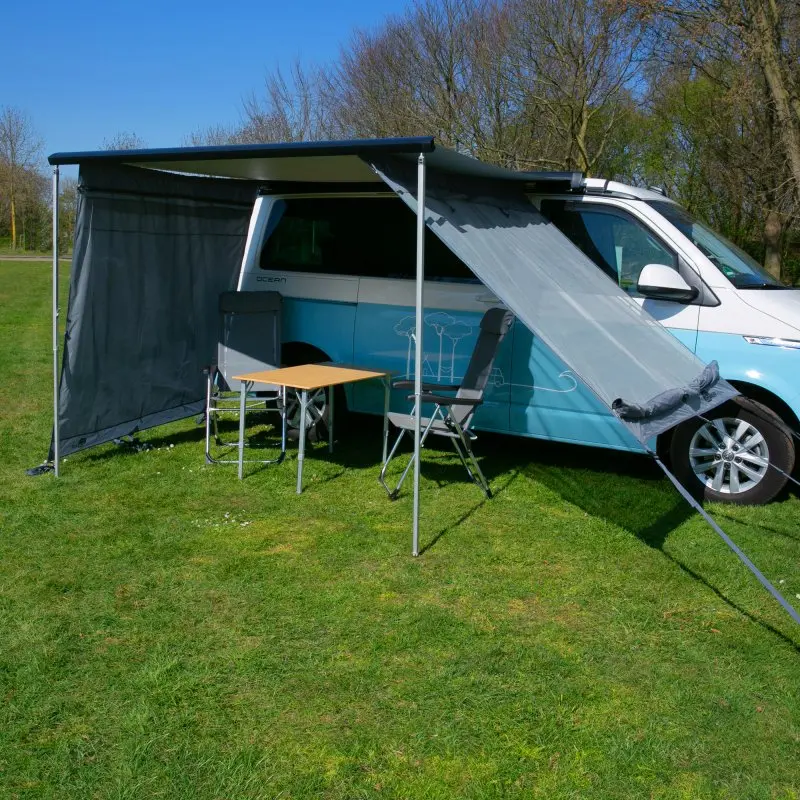 your GEAR Campingbus Sun + Rain Blocker side walls mounted on a van awning, showing two side panels (180 x 190 cm) for sun and rain protection, with table and chairs underneath, outdoor setting.
