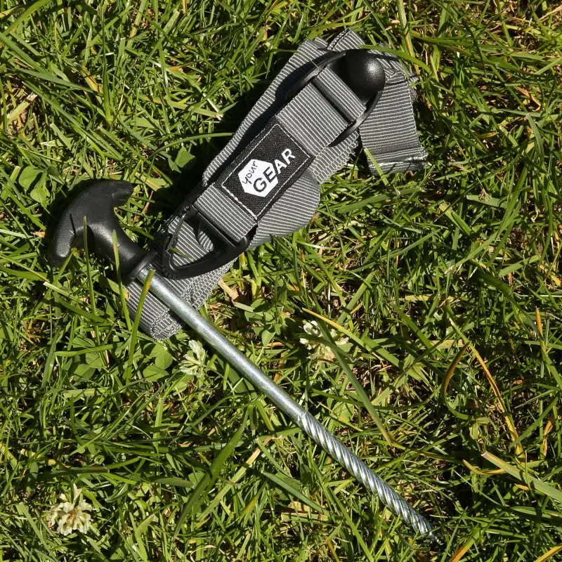 Set de haubanage your GEAR : sangle grise en polyester avec boucle réglable, mousqueton noir et piquet de sol en métal, posé sur l’herbe. Accessoire pour tente, camping et auvent.
