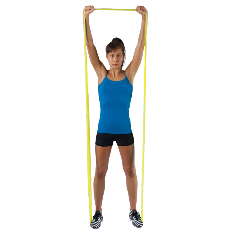 Woman exercising with a yellow Spirit Fitness Band 9 kg, latex-free, 183x10 cm, holding the resistance band overhead while standing, demonstrating its use for strength and rehabilitation training.