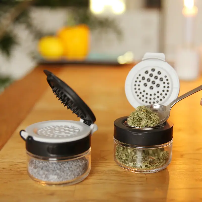 Two your GEAR Double-Flip spice jars made of clear glass with black and white dual-flip lids, filled with herbs and spices, shown open on a wooden kitchen table for easy seasoning and storage.