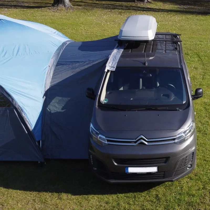 your GEAR Bergamo 430 Bus Connector attached between a gray van and a blue event pavilion, showing the weather-protected tunnel connection for camping, with visible zippers and secure fit on grass.