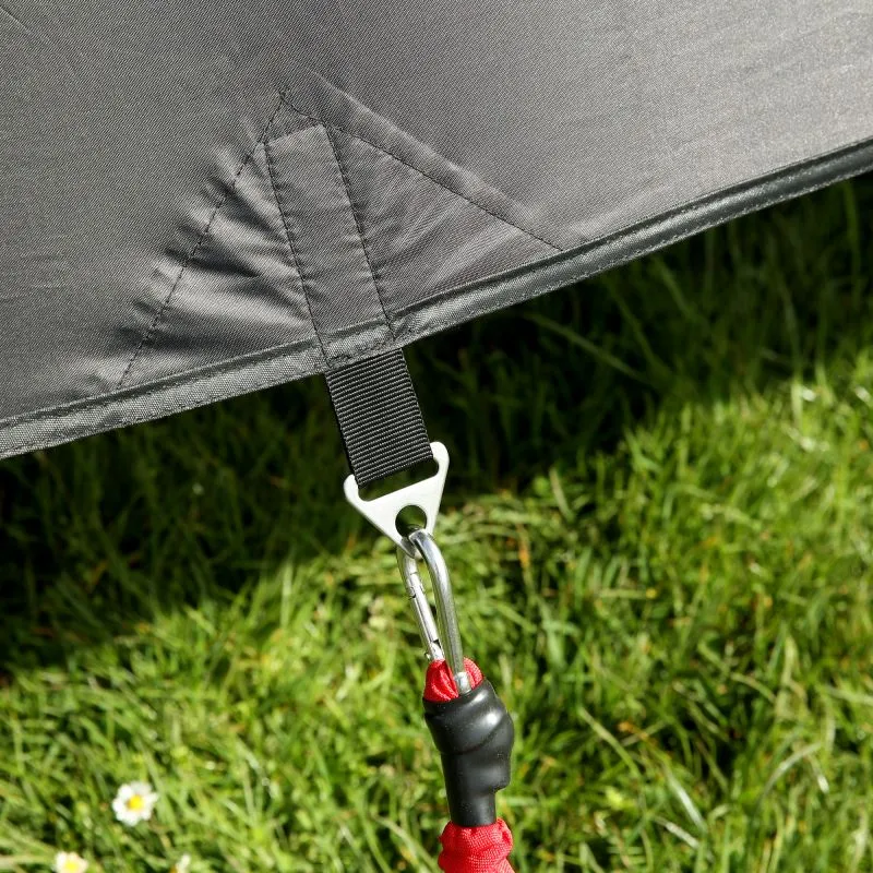 Détail de la face avant du multicover your GEAR Phoenix en tissu gris argenté, couture renforcée, sangle noire avec mousqueton métallique attaché à une cordelette rouge, posé au-dessus d’une pelouse verte.