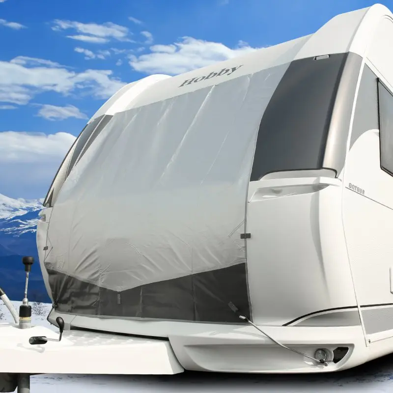 Caravana blanca con Bugschutzplane Thermo your GEAR de 165 x 160 cm instalada en el frontal, protege contra frío y calor, fondo de montaña, ideal para invierno y verano, fijación segura visible.