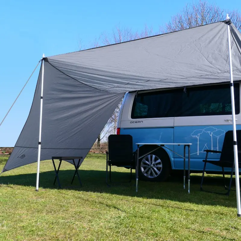 your GEAR Tropea bus awning with UV 50+ protection, gray tarp with two side walls, set up on a van with aluminum poles; outdoor camping setup with chairs and table underneath.