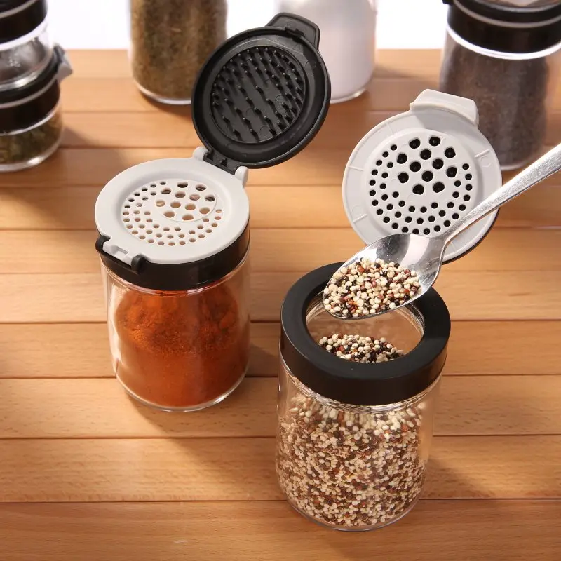Two your GEAR Double-Flip spice jars made of glass, each with a black and gray dual-function flip lid, filled with spices, shown on a wooden surface, demonstrating easy pouring and sprinkling.