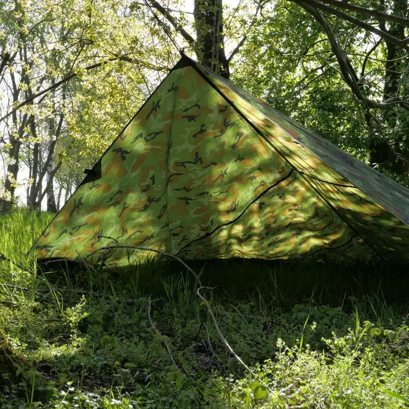 Tarp yourGEAR Bushcraft 3x3 m camouflage, installé comme abri sous les arbres. Toile imperméable et robuste pour camping ou survie, protection UV 50+, adaptée à l'extérieur, visible dans un environnement forestier.
