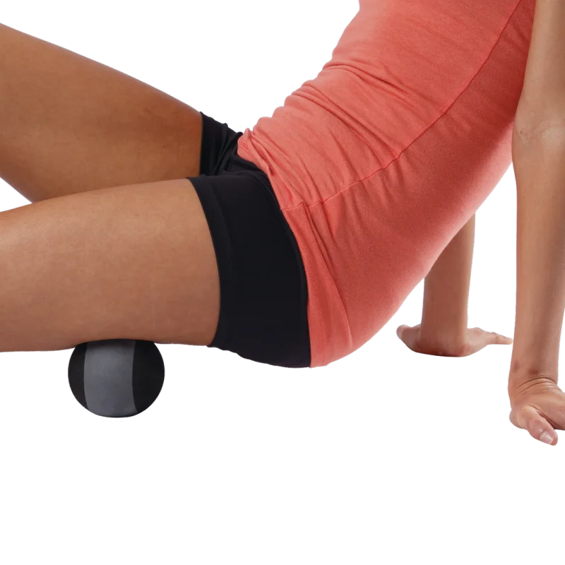 Woman using a Spirit Massageball Ø 8.9 cm under her thigh for fascia and trigger point massage; black and grey ball shown in use for muscle recovery and self-massage.