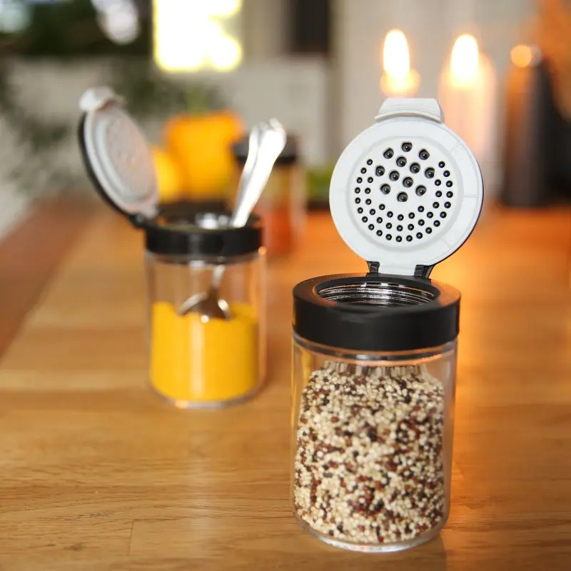 your GEAR Double-Flip spice jars with black lids, open dual-function lids for pouring or sprinkling, filled with spices, displayed on a wooden kitchen table, ideal for organized spice storage.